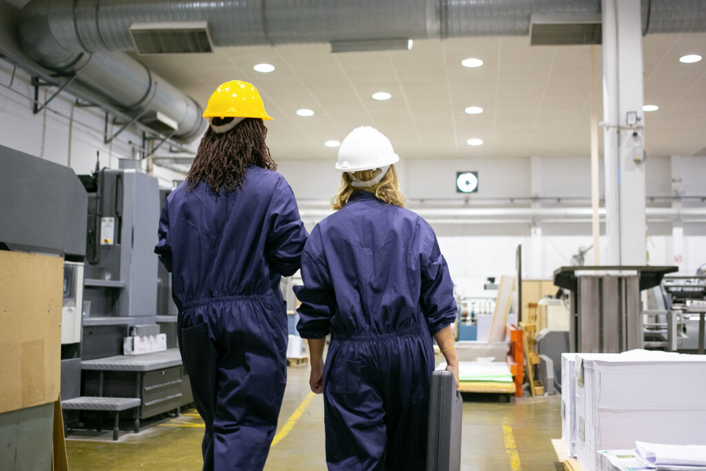 Deux ouvrières en casque et combinaison de travail marchant dans un atelier industriel : prévention des risques et santé au travail